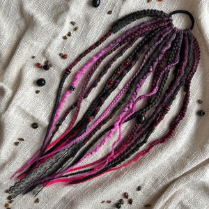 May include: A collection of black, pink, and red dreadlocks, fastened with a black elastic. The dreadlocks are decorated with small beads and other embellishments, displayed on a textured, off-white background.