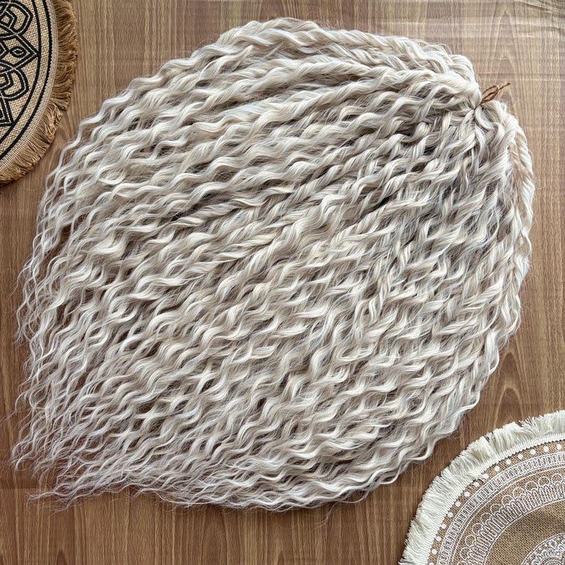 Puede incluir: Un paquete de extensiones de cabello largo, blanco y rizado. El cabello est&aacute; dispuesto en una pila suelta y ondulada.