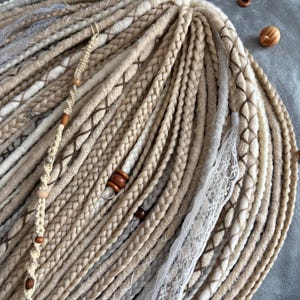 Könnte beinhalten: Nahaufnahme einer Sammlung von blonden und hellbraunen Dreadlocks. Einige Dreadlocks sind mit Holzperlen und dekorativer Spitze verziert. Die Dreadlocks sind fächerförmig angeordnet und zeigen verschiedene Texturen und Farbtöne.
