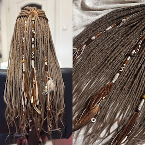 Può includere: Dreadlock e trecce sintetiche color marrone chiaro con accenti decorativi tra cui perline di legno, avvolgimenti in filo bianco e marrone, ciondoli metallici e piume naturali. Le extension sono acconciate in una mezza coda.