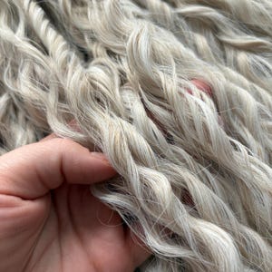 Puede incluir: Primer plano de un paquete de extensiones de cabello ondulado gris claro. El cabello es suave y sedoso, con un aspecto natural ligeramente texturizado.