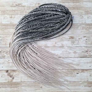 May include: A bundle of braided synthetic hair extensions in a gradient of colors. The top section is dark gray, transitioning to lighter gray and then a pale blonde at the ends. The braids are neatly arranged.
