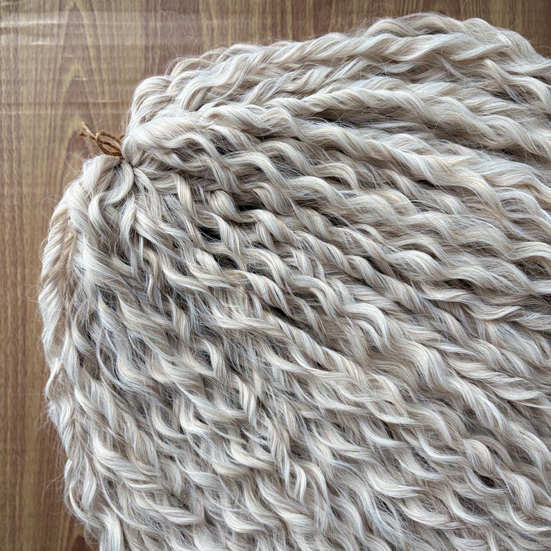 Puede incluir: Primer plano de un paquete de extensiones de cabello ondulado, rubio claro y blanco. El cabello est&aacute; dispuesto en un estilo suelto y fluido.