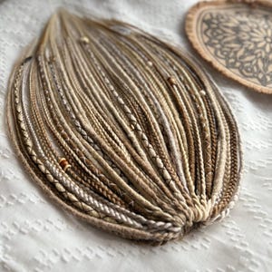 Könnte beinhalten: Ein Bündel brauner, beigefarbener und weißer synthetischer Haarverlängerungen mit braunen Perlen. Das Haar ist in einer kreisförmigen Form angeordnet.