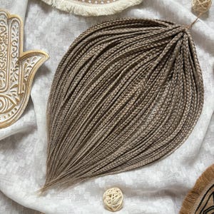 May include: A bundle of light brown synthetic hair braids, arranged in a teardrop shape. The braids are tightly woven and have a natural, textured appearance. The background is a white fabric with decorative elements.