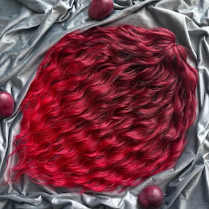 May include: A vibrant, wavy wig with a gradient of red hues, transitioning from a deep burgundy at the roots to a bright, fiery red at the ends. The wig is styled on a silver fabric, with three red apples scattered around.