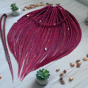 Könnte beinhalten: Ein Set aus dunkelburgunderfarbenen synthetischen Dreadlocks und Zöpfen mit goldfarbenen Drahtwicklungen und kleinen weißen Muschelakzenten. Die Haarverlängerungen sind fächerförmig angeordnet, mit einer langen geflochtenen Strähne links.