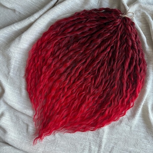 Op de afbeelding: Een bundel rode en donkerrode synthetische hairextensions, gerangschikt in een golvende, gelaagde stijl. Het haar is aan de bovenkant samengebonden met een natuurlijk gekleurd touwtje. De extensions worden gepresenteerd op een neutraal gekleurde stof.