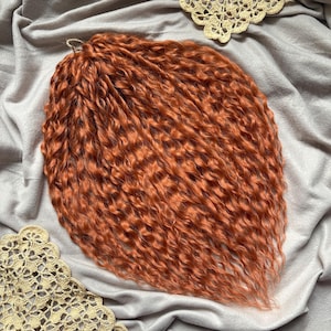 May include: A mass of vibrant, reddish-orange curly hair extensions. The hair is tightly curled and appears to be synthetic. A metal loop is attached at the top. The hair is displayed on a gray fabric surface with decorative lace.