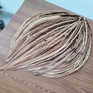 May include: A set of brown, beige, and white synthetic hair extensions with beads and lace accents. The extensions are arranged in a fan shape on a wooden surface.