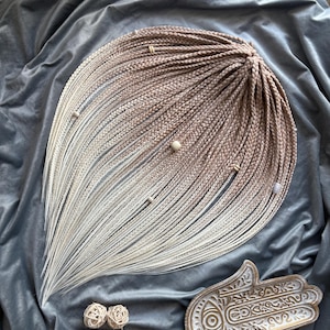 Peut inclure: Un ensemble d'extensions capillaires synthétiques composé de nombreuses fines tresses avec un dégradé de couleur allant d'un brun moyen chaud aux racines vers un blanc crème clair aux pointes, agrémenté de perles en bois et d'anneaux.