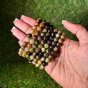May include: A stack of green, brown and black gemstone bracelets. The bracelets are made of round beads and are stacked on top of each other.