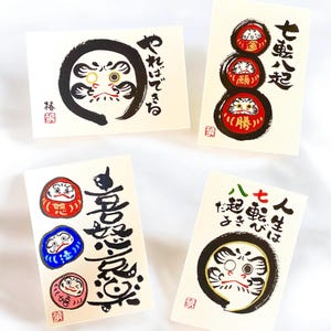 May include: Four rectangular cards with Japanese calligraphy and Daruma doll illustrations. The cards feature black, red, blue, and pink Daruma dolls, with Japanese text in black ink. The cards are on a white background.