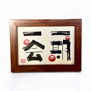 以下が含まれることがあります： クリーム色の背景に黒インクで描かれた日本の書道が特徴の額装アート。木製のフレームは濃い茶色です。アートワークには赤いダルマ人形と赤いスタンプが含まれています。