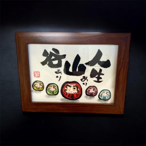 May include: A framed artwork featuring Japanese calligraphy and Daruma dolls. The calligraphy is in black ink, and the Daruma dolls are in various colors, including red, green, and yellow. The frame is made of dark wood.