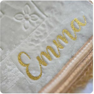 Könnte beinhalten: Eine weiße Plüschdecke mit einem goldenen gestickten Namen "Emma" darauf. Die Decke hat einen beigefarbenen Saum an den Rändern.