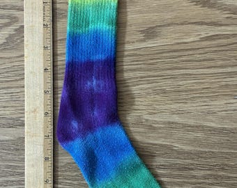 May include: A ribbed crew sock featuring a tie-dye rainbow pattern with horizontal bands of red, orange, yellow, green, blue, and purple. The sock is positioned next to a wooden ruler showing a length of approximately 12 inches from top to toe.
