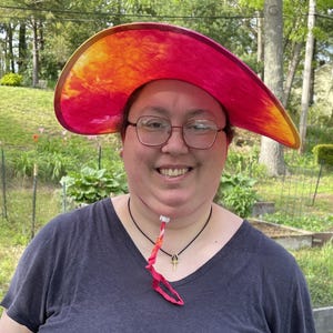 May include: A wide-brimmed sun hat featuring a vibrant tie-dye pattern in shades of bright pink, orange, and yellow. The hat includes a matching tie-dye chin strap with an adjustable white plastic toggle to secure the fit under the chin.