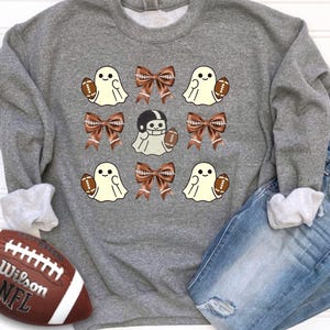 May include: A gray sweatshirt featuring a repeating pattern of cartoon ghosts holding footballs and brown bows. One ghost wears a football helmet. A brown and white football and a pair of blue jeans are also in the image.