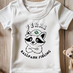 Op de afbeelding: Wit T-shirt met een cartoon wasbeer met een honkbalpet. De wasbeer heeft zwart-witte markeringen en gekruiste armen. De tekst "FERAL BALLPARK SIBLING" is afgedrukt.