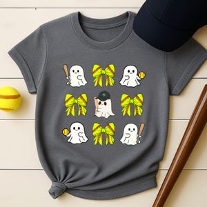May include: Gray t-shirt featuring a playful design of cartoon ghosts with baseball bats, softballs, and bows. A baseball cap and a softball are also visible. The shirt is a comfortable, casual wear item.