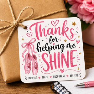 Peut inclure: Une étiquette cadeau avec les mots "Thanks for helping me SHINE" en écriture rose. L'étiquette présente des chaussons de ballet roses et des cœurs et étoiles décoratifs. L'étiquette est attachée à un cadeau emballé marron avec de la ficelle.