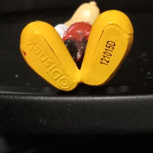 Może przedstawiać: Ż&oacute;łta plastikowa figurka Myszki Miki leżąca na plecach. Sp&oacute;d st&oacute;p jest oznaczony "&copy;Disney" i "121015DC".