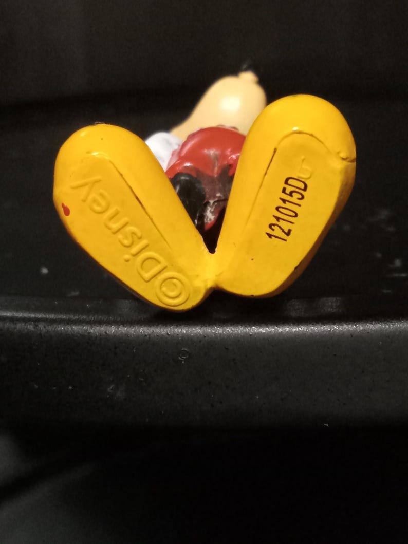 Może przedstawiać: Ż&oacute;łta plastikowa figurka Myszki Miki leżąca na plecach. Sp&oacute;d st&oacute;p jest oznaczony "&copy;Disney" i "121015DC".