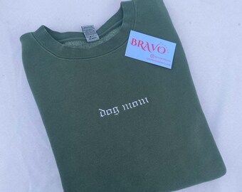 Könnte beinhalten: Ein dunkelgrüner Pullover mit dem Schriftzug "dog mom" in weiß auf der Vorderseite gestickt. Der Pullover ist gefaltet und liegt flach auf einer weißen Oberfläche.