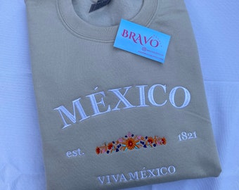 May include: Tan crewneck sweatshirt with white embroidered text "MÉXICO" and "VIVA MÉXICO". Features a floral design and the text "est. 1821". A small tag with the word "BRAVO" is attached.