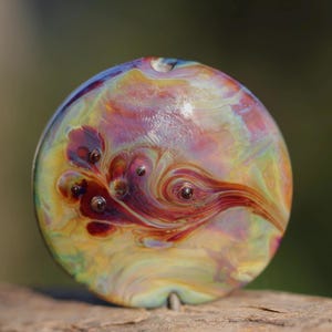 May include: A circular glass pendant featuring an iridescent finish with swirling patterns of deep red, amber, and gold. The surface displays a smooth, polished texture with small, raised dark red circular inclusions embedded within the glass.