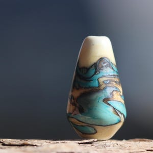 May include: A teardrop-shaped pendant bead featuring a smooth, polished surface with a swirling pattern of cream, dark brown, and vibrant turquoise blue colors. The bead has a small hole at the top for stringing, with a glossy finish.