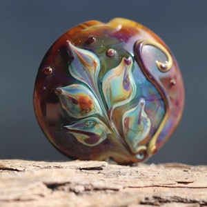 May include: A round, polished glass bead featuring an iridescent, multi-colored floral design with swirling teal, purple, and cream petals set against a deep, translucent amber background with small, raised glass dots.