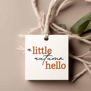 May include: A square white paper tag with a hole at the top, threaded with natural tan twine. The text "a little autumn hello" is printed on the front in a combination of burnt orange block letters and black cursive script lettering.