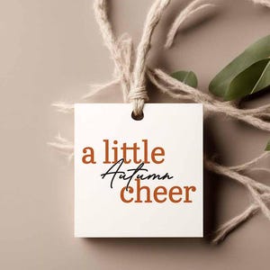 Könnte beinhalten: Ein quadratisches weißes Papieretikett mit einem Loch an der Oberseite, durch das eine naturfarbene Schnur gezogen ist. Der Text "a little Autumn cheer" ist auf der Vorderseite aufgedruckt, wobei "a little" und "cheer" in Rostorange und "Autumn" in schwarzer Schreibschrift gehalten sind.