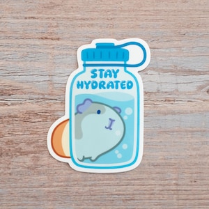 Peut inclure: Autocollant représentant un cobaye de dessin animé dans une bouteille d'eau. La bouteille bleue porte l'inscription "STAY HYDRATED". Le cobaye est bleu clair et gris. L'autocollant a une bordure blanche et un accent orange.