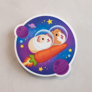 May include: Two guinea pigs wearing astronaut helmets are riding a carrot rocket through space. The background is a purple galaxy with stars and planets.