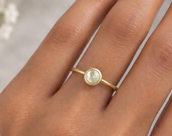 May include: A gold ring featuring a single round, bezel-set white gemstone. The band is thin and polished, showcasing a minimalist design. The ring is worn on a finger, highlighting the circular stone setting and the smooth, metallic finish of the band.