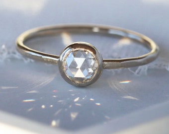 May include: A delicate, gold-toned ring featuring a clear, faceted gemstone. The gemstone is set in a bezel setting, and the band is thin and polished. The ring is displayed on a light-colored surface, with soft shadows.