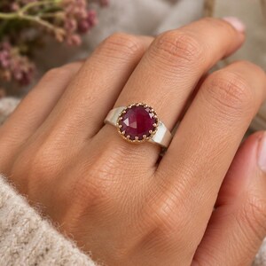 May include: A ring featuring a round, faceted deep red gemstone set in a gold-toned scalloped bezel, mounted on a smooth, wide silver-toned band. The stone has a textured, reflective surface and is positioned centrally on the finger.