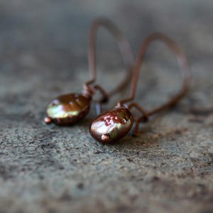 May include: A pair of copper-colored earrings with teardrop-shaped, iridescent, brown-toned beads. The earrings have a simple hook design. The background is a textured, gray surface. The earrings are a jewelry item.