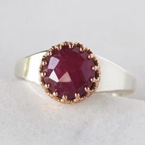 May include: A silver ring featuring a large, faceted, deep red gemstone. The gemstone is set in a gold-coloured, crown-like bezel. The ring band is a simple, polished silver.
