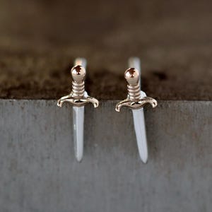 May include: A pair of silver-colored sword-shaped earrings with gold-colored accents. The earrings have a detailed hilt and a pointed blade. The background is a blurred, neutral tone.