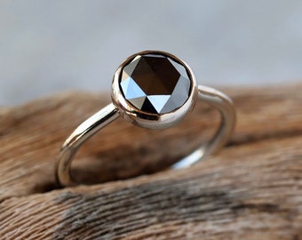 May include: A silver ring featuring a large, faceted black gemstone. The ring has a simple, elegant design with a polished band and a bezel setting that holds the dark stone. The ring is displayed on a piece of wood.