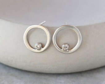 May include: A pair of silver stud earrings. Each earring has a small, clear gemstone set within a circular, open design. The earrings have a minimalist, modern aesthetic, and are displayed on a light grey surface.