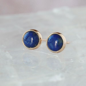 May include: A pair of gold stud earrings featuring round, deep blue lapis lazuli stones. The earrings have a simple, elegant design, with the gold setting encircling the blue stone. The studs are displayed on a white, textured surface.