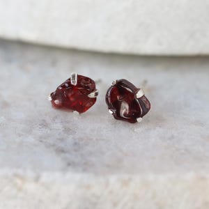 May include: A pair of raw garnet stud earrings. The earrings feature deep red, irregularly shaped gemstones set in silver-colored metal. The earrings are displayed on a white and gray marble surface.