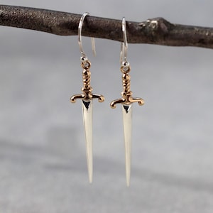 May include: Silver-tone sword earrings with gold-colored accents. The earrings are hanging from a branch. The swords have a detailed hilt and a pointed blade. The earrings are a unique accessory.