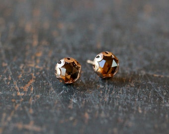 May include: Two gold-colored stud earrings with faceted, brown gemstones. The earrings have a round shape with decorative cutouts around the edges. The studs are set against a dark, textured background.