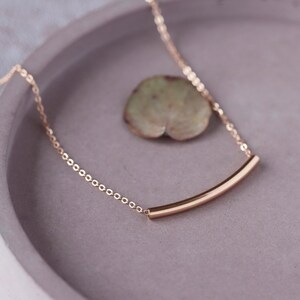 May include: A delicate gold-colored necklace with a curved bar pendant. The necklace chain is made of small, connected links. The pendant is a smooth, curved bar. The necklace rests on a gray surface with a green leaf.